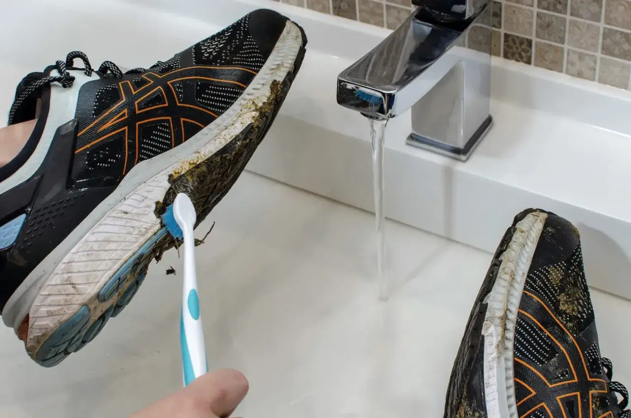 Come lavare le scarpe da running: una mano usa uno spazzolino per pulire il fango dalla suola di una scarpa da corsa sotto un rubinetto aperto.