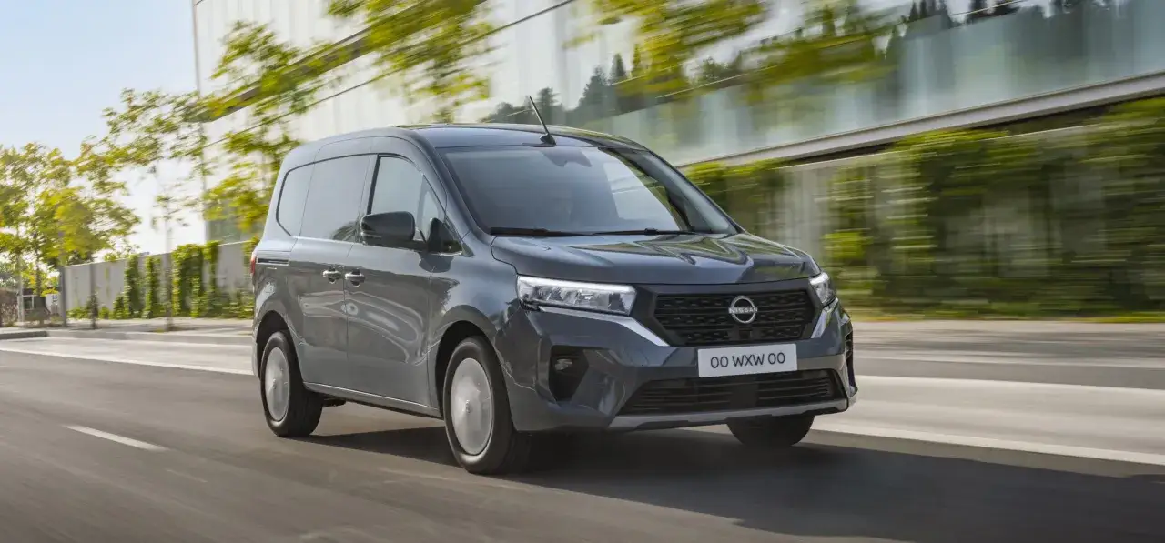 Szary Nissan Townstar pędzi po drodze, otoczony zielenią i nowoczesną architekturą.