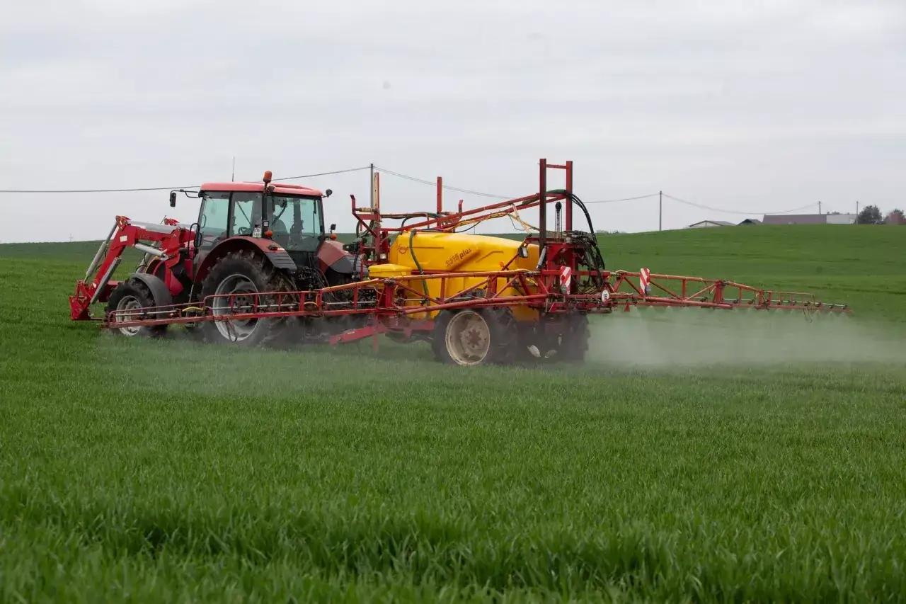 Czerwony traktor z żółtym opryskiwaczem, model halvetic, pracuje na zielonym polu, rozpylając środek ochrony roślin.