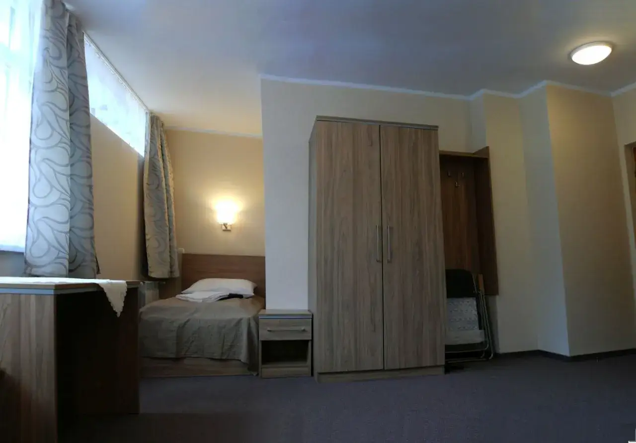 Pokój w sanatorium Leśnik w Sopocie: łóżko, szafa, biurko i okno z zasłonami.