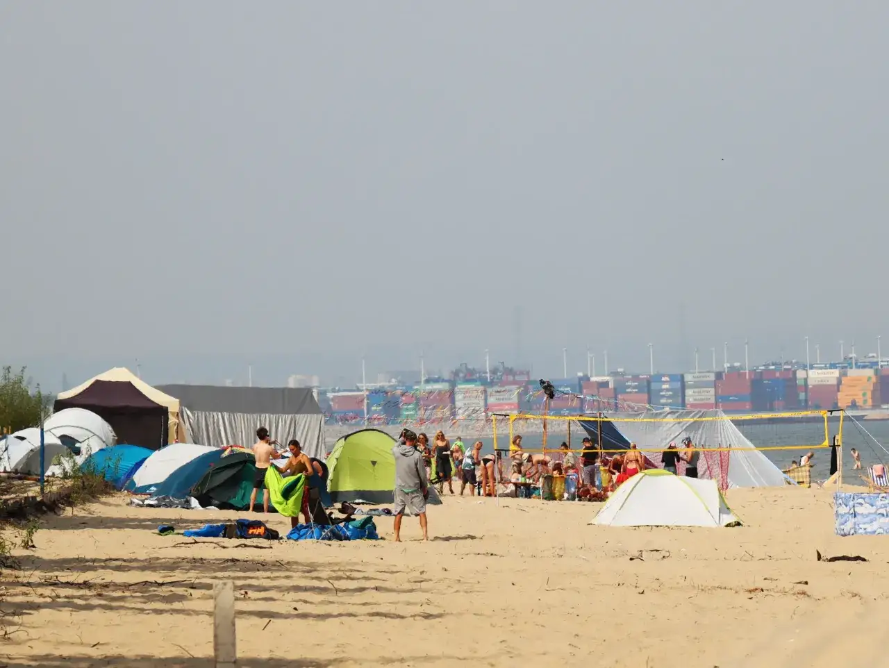 Ludzie rozbijają namioty na plaży, tworząc tymczasowe obozowisko. W tle widać port z kontenerowcami.