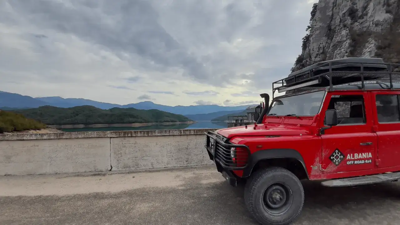 Czerwony Land Rover na tle górskiego jeziora. Chmurne niebo, typowa albania pogoda styczeń.