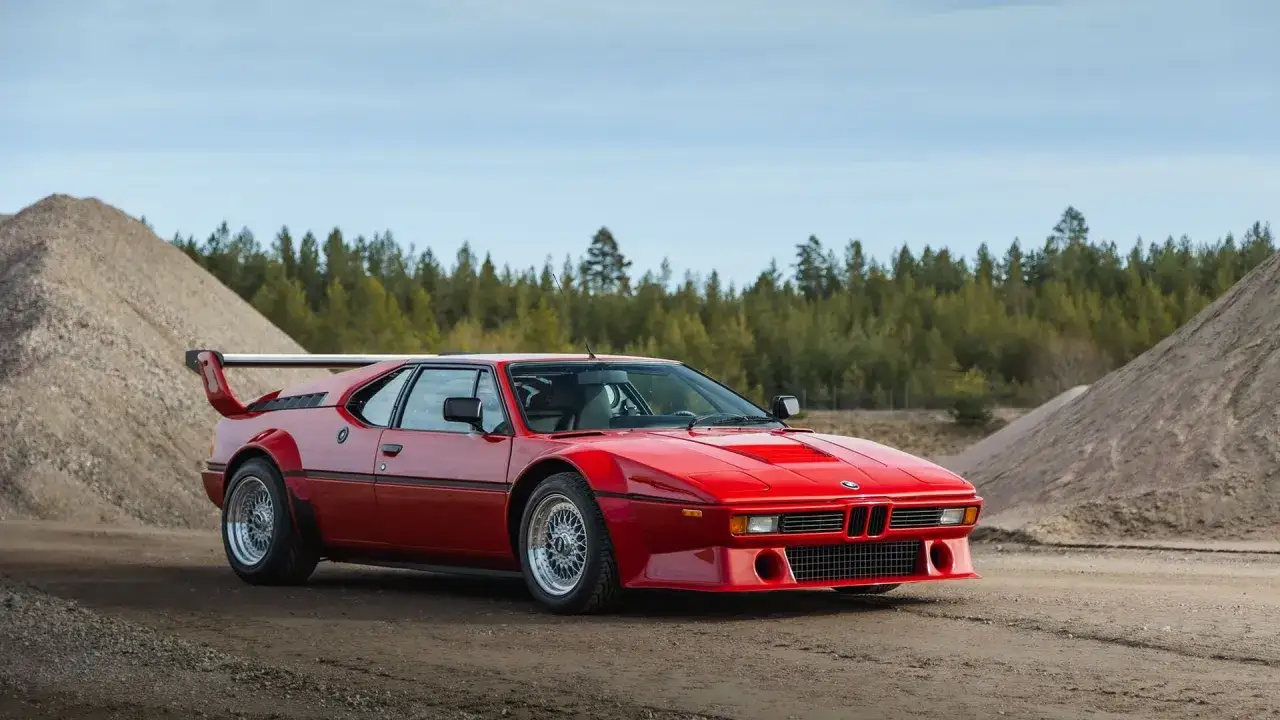 El ic&oacute;nico m1 coche rojo, con su aler&oacute;n distintivo, descansa en un camino de tierra, rodeado de montones de grava y un bosque de pinos.