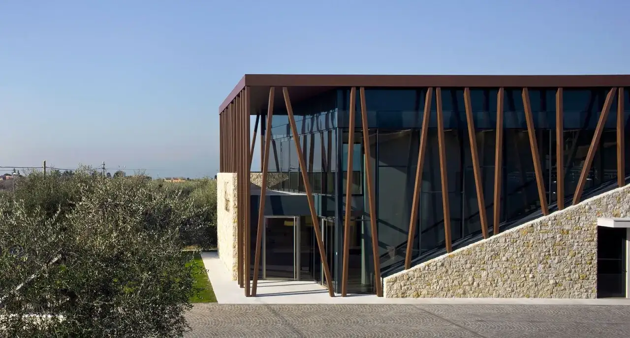 Edificio moderno con facciata in pietra e vetro, circondato da ulivi. Un'architettura che evoca i **valetti** di un vigneto a **Bardolino**.