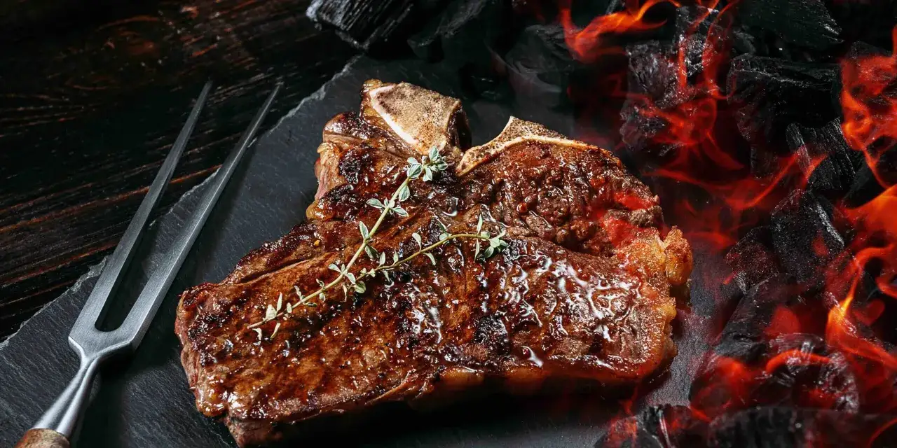 Un jugoso chuleton a la brasa, con romero fresco, listo para ser servido junto a brasas ardientes.