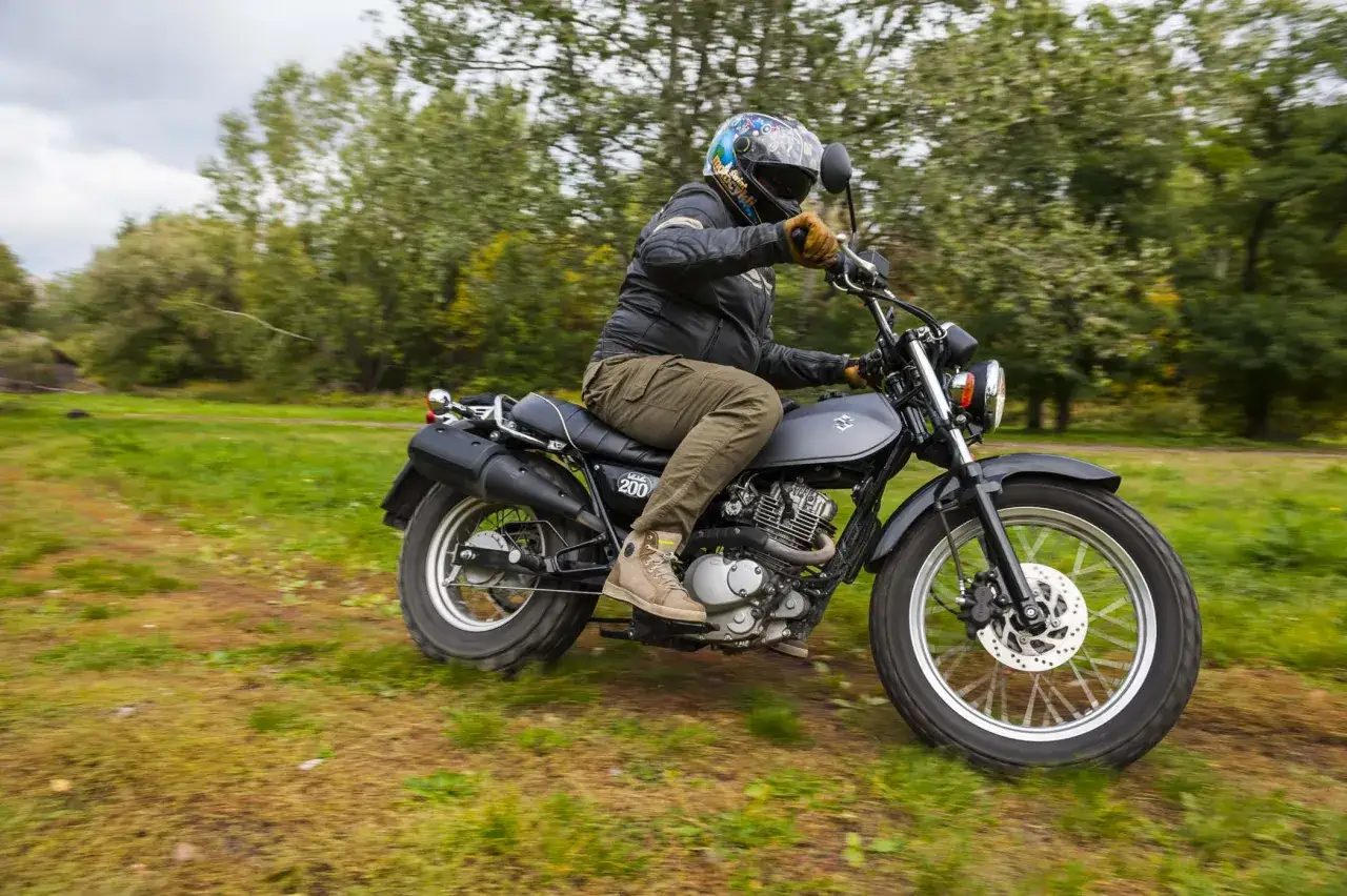 Motocyklista na Suzuki Van Van 200 przemierza zieloną łąkę wśród drzew.