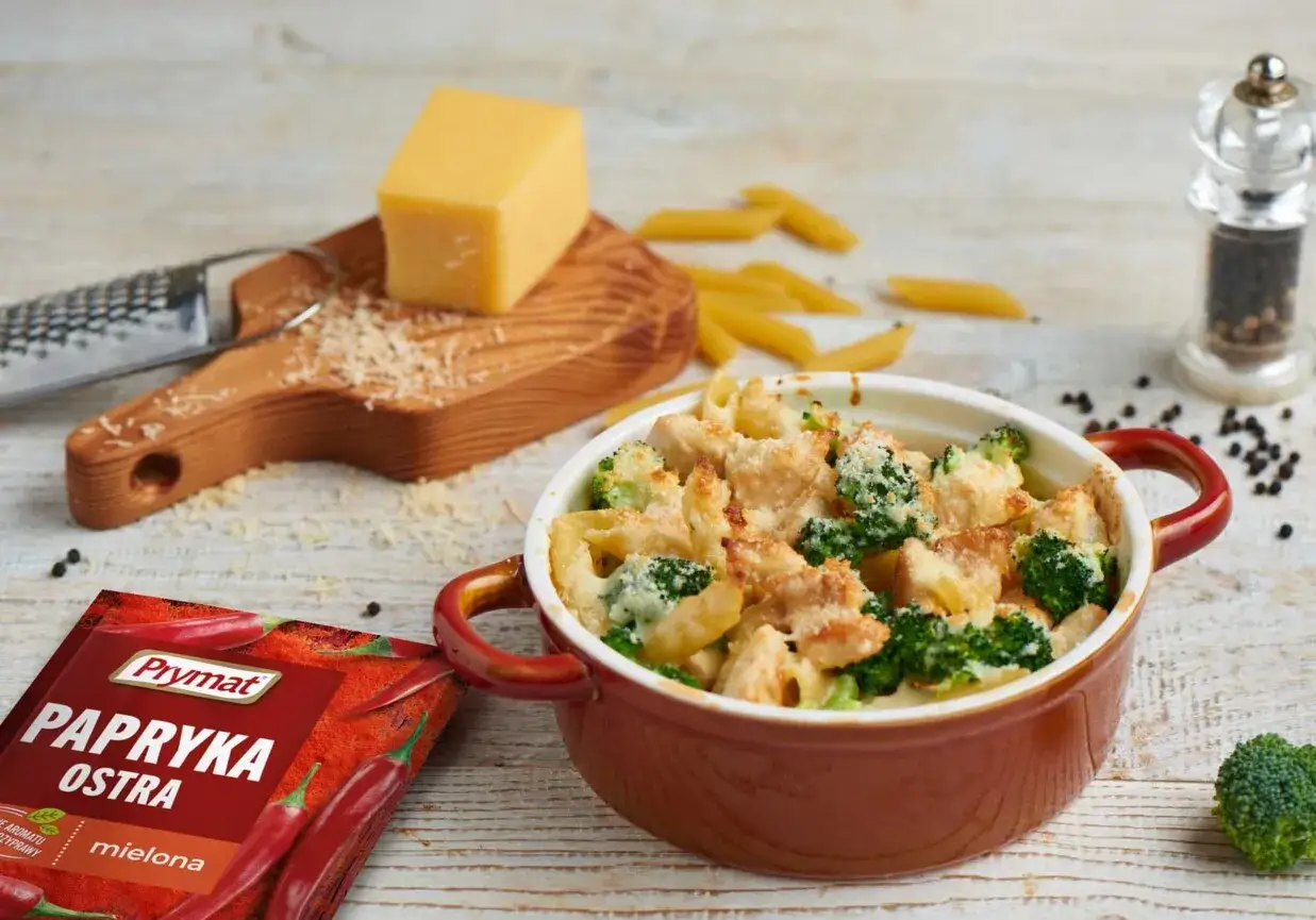 Pyszna zapiekanka z makaronem i kurczakiem i brokułem, posypana parmezanem. Obok kawałek sera, przyprawy i surowy brokuł.