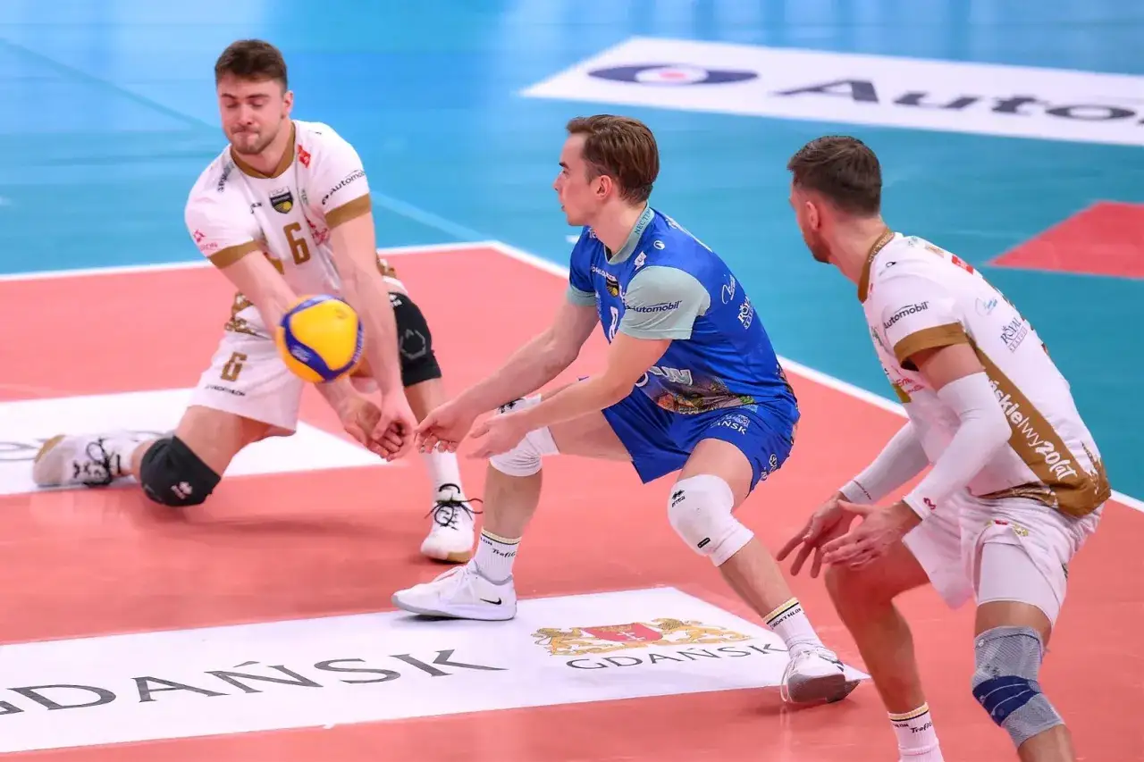 Rafał Sobański w akcji, przyjmuje piłkę siatkową. Gracze w białych i niebieskich strojach walczą o każdy punkt.