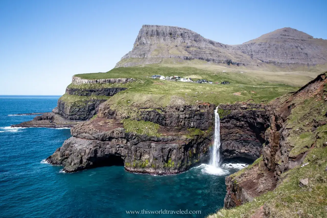 Cascata che si getta nell'oceano, con scogliere verdi e un villaggio in lontananza. Un vero spettacolo **far oer**.