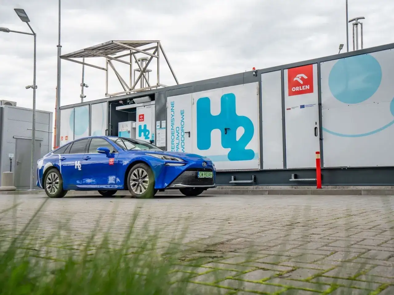 Niebieski samochód na wodór zaparkowany przy stacji paliw Orlen. H2 symbolizuje przyszłość motoryzacji.