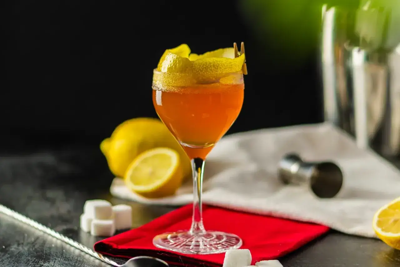 Elegantes Cognac-Cocktail mit Zitronenzeste, garniert mit einem kleinen W&auml;scheklammer-Detail. Zitronen und Zuckerw&uuml;rfel deuten auf die Zubereitung hin.
