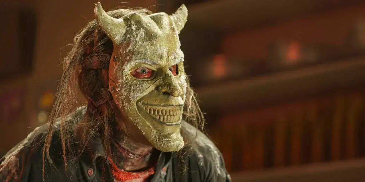 Un volto terrificante con maschera da demone, capelli lunghi e occhi rossi. La maschera, che sembra il seme della follia, è cosparsa di polvere bianca.