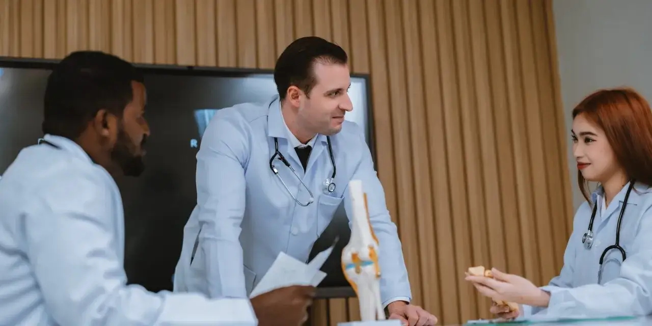Medici discutono un caso, con un modello di ginocchio e una radiografia. Uno è un fisiatra o ortopedico.