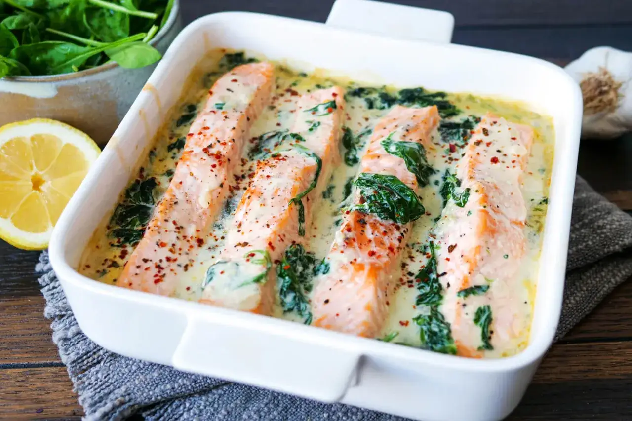Vier saftige Lachsfilets in cremiger Spinatsauce, bestreut mit Gew&uuml;rzen, in einer wei&szlig;en Auflaufform. Daneben liegen frischer Spinat, eine Zitronenh&auml;lfte und Knoblauch.