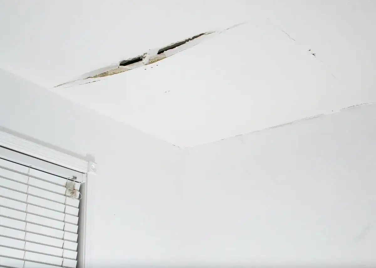 Fissure b&eacute;ante dans un plafond en placo, n&eacute;cessitant une r&eacute;paration. Un coin de fen&ecirc;tre avec stores est visible.