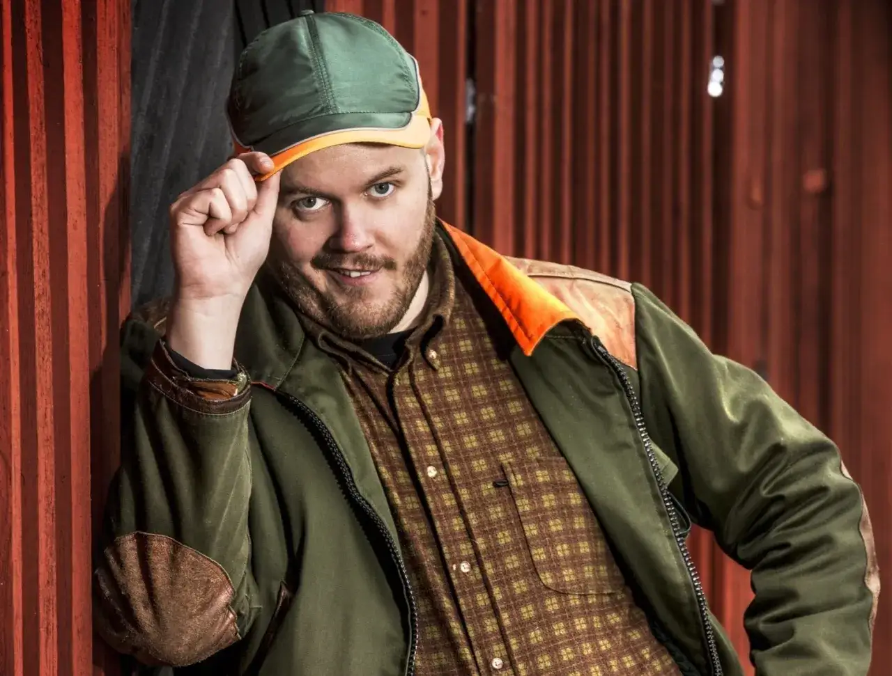 En leende man med skägg och keps, klädd i en rutig skjorta och jacka. Kanske en scen från leif och billy film?