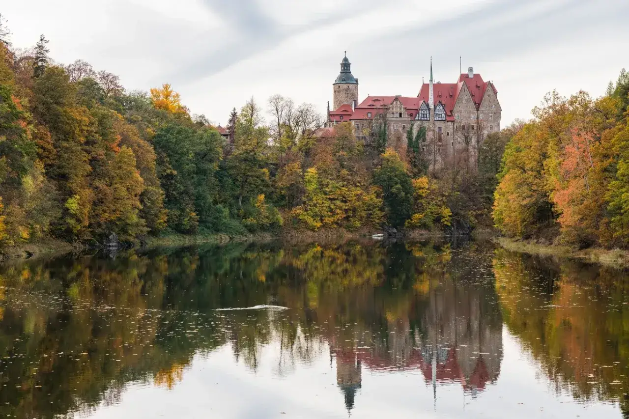 Zamek Czocha, gdzie jest, otoczony jesiennymi drzewami, odbija się w spokojnej tafli jeziora.