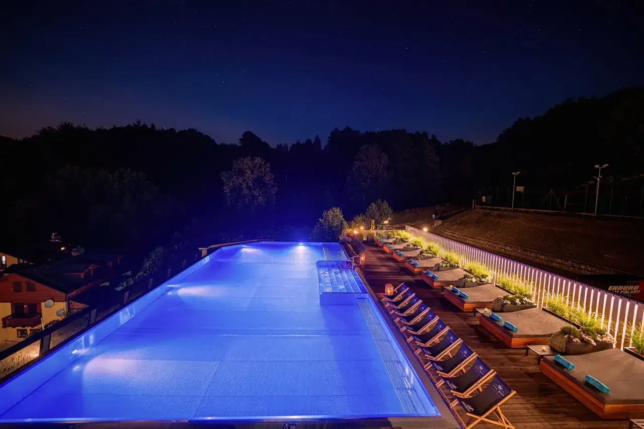 Nocny basen z niebieskim podświetleniem i leżakami, idealny dla hotelu z widokiem na góry. Gwiaździste niebo nad lasem.
