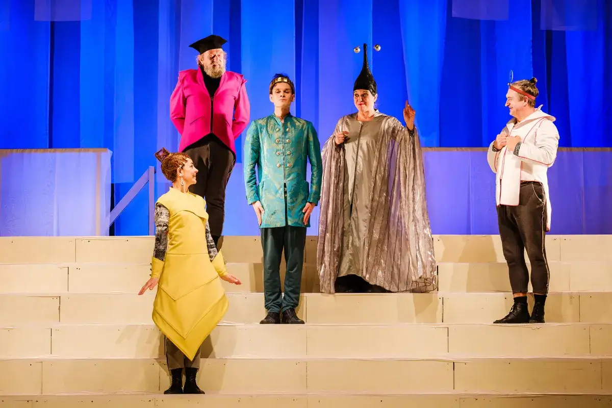 Aktorzy w barwnych strojach na scenie, zapowiedź spektaklu z repertuaru teatru lalkowego.