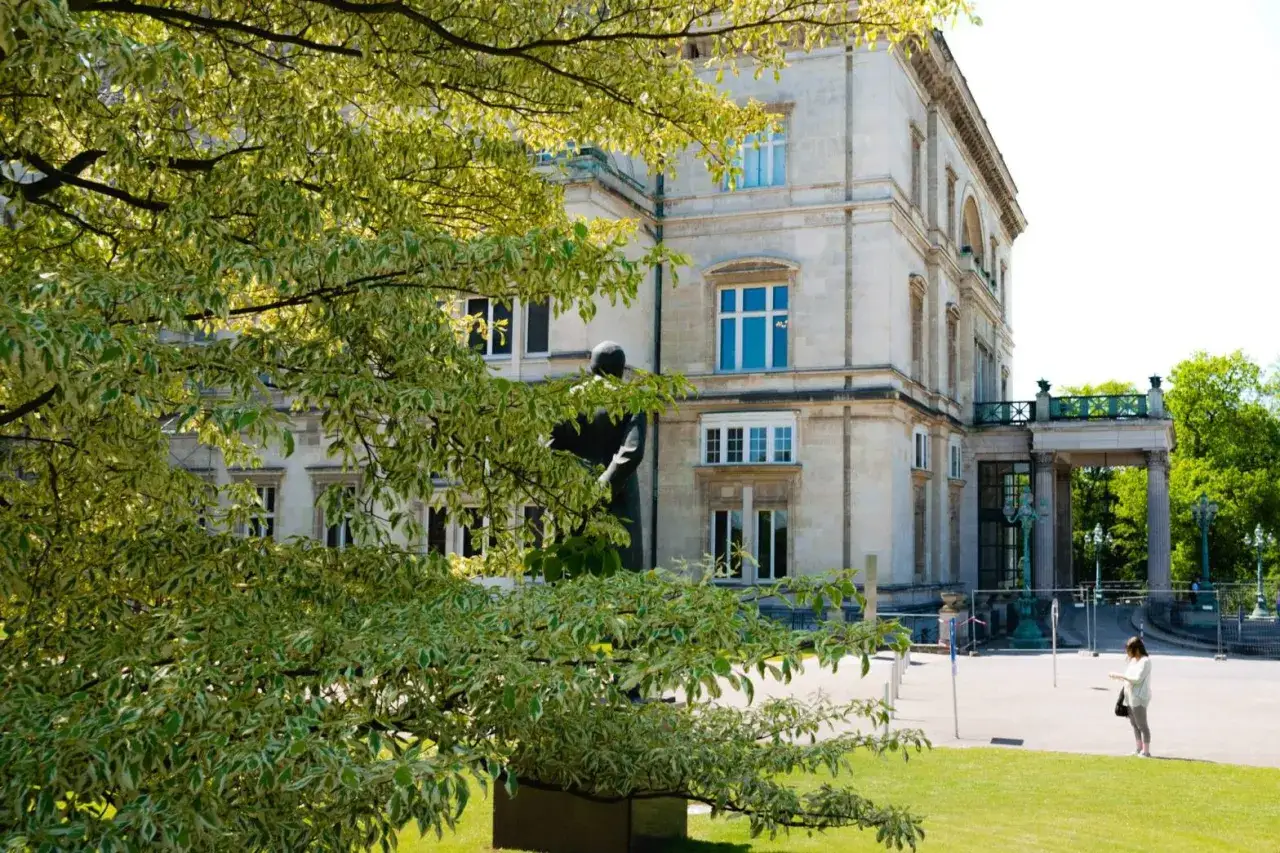 Pr&auml;chtige Krupp Villa mit Statue im Vordergrund, umgeben von &uuml;ppigem Gr&uuml;n und einem Baum mit bunten Bl&auml;ttern.
