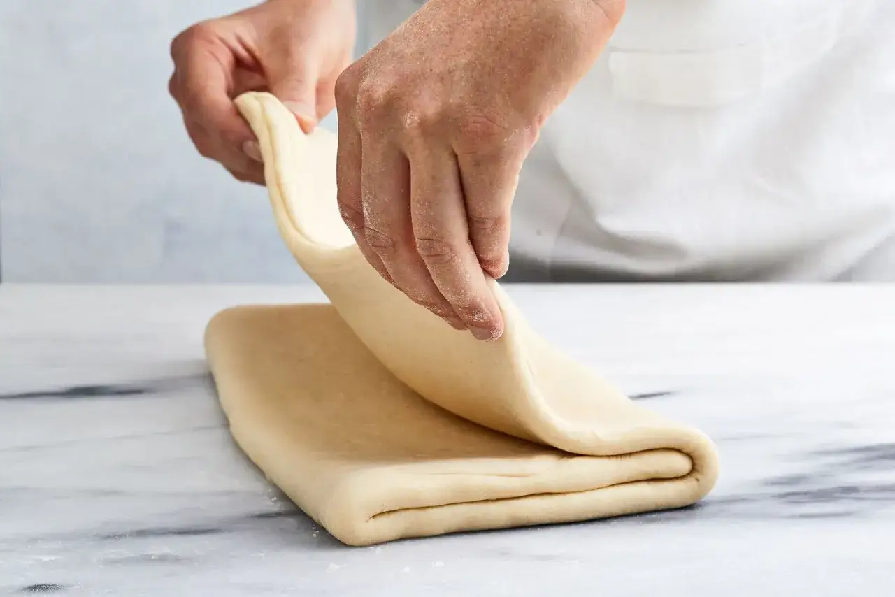 Manos pliegan masa hojaldrada, un paso clave en los ingredientes de croissant.