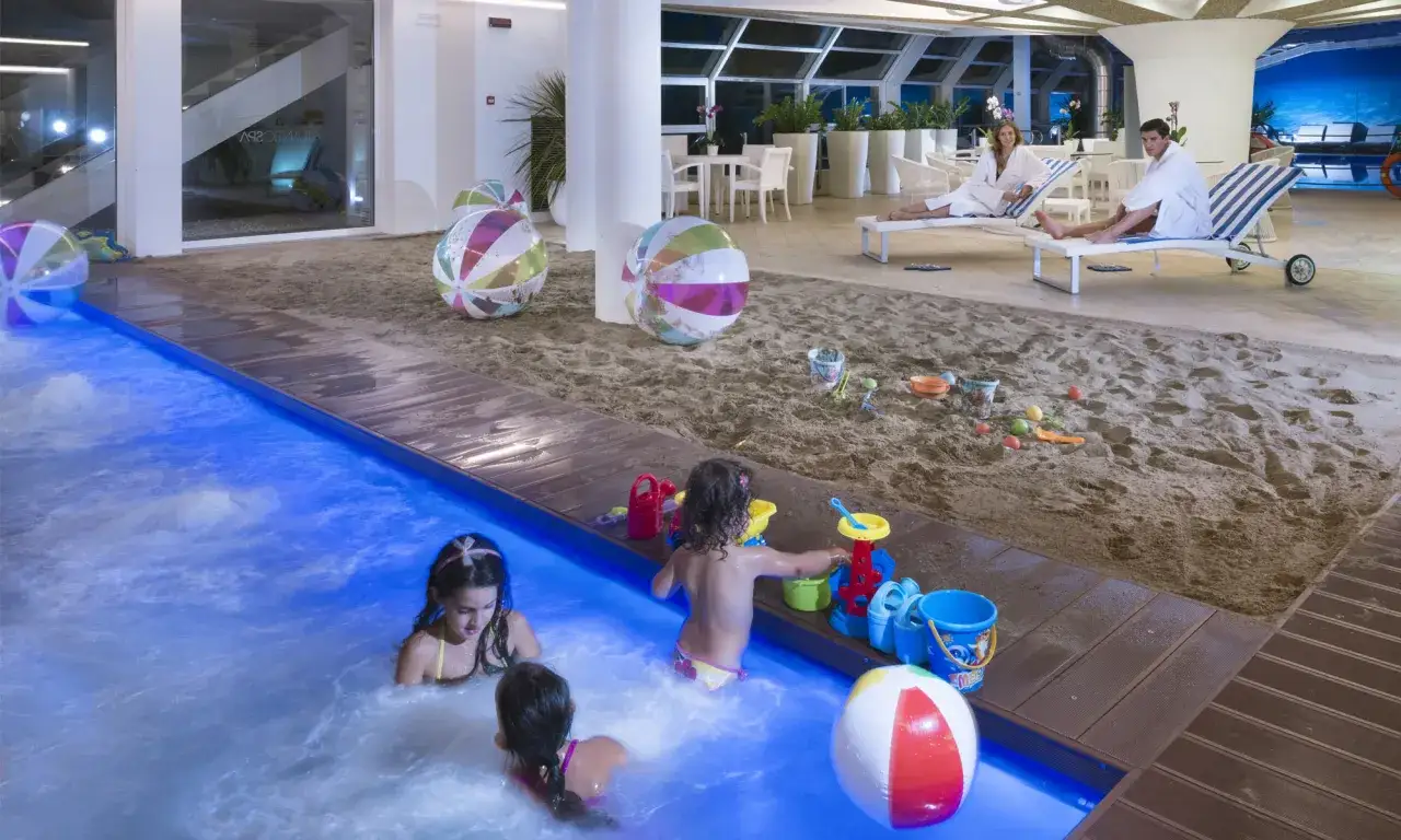 Bambini giocano in piscina mentre adulti si rilassano sulla sabbia in questi bagni Riccione con piscina.