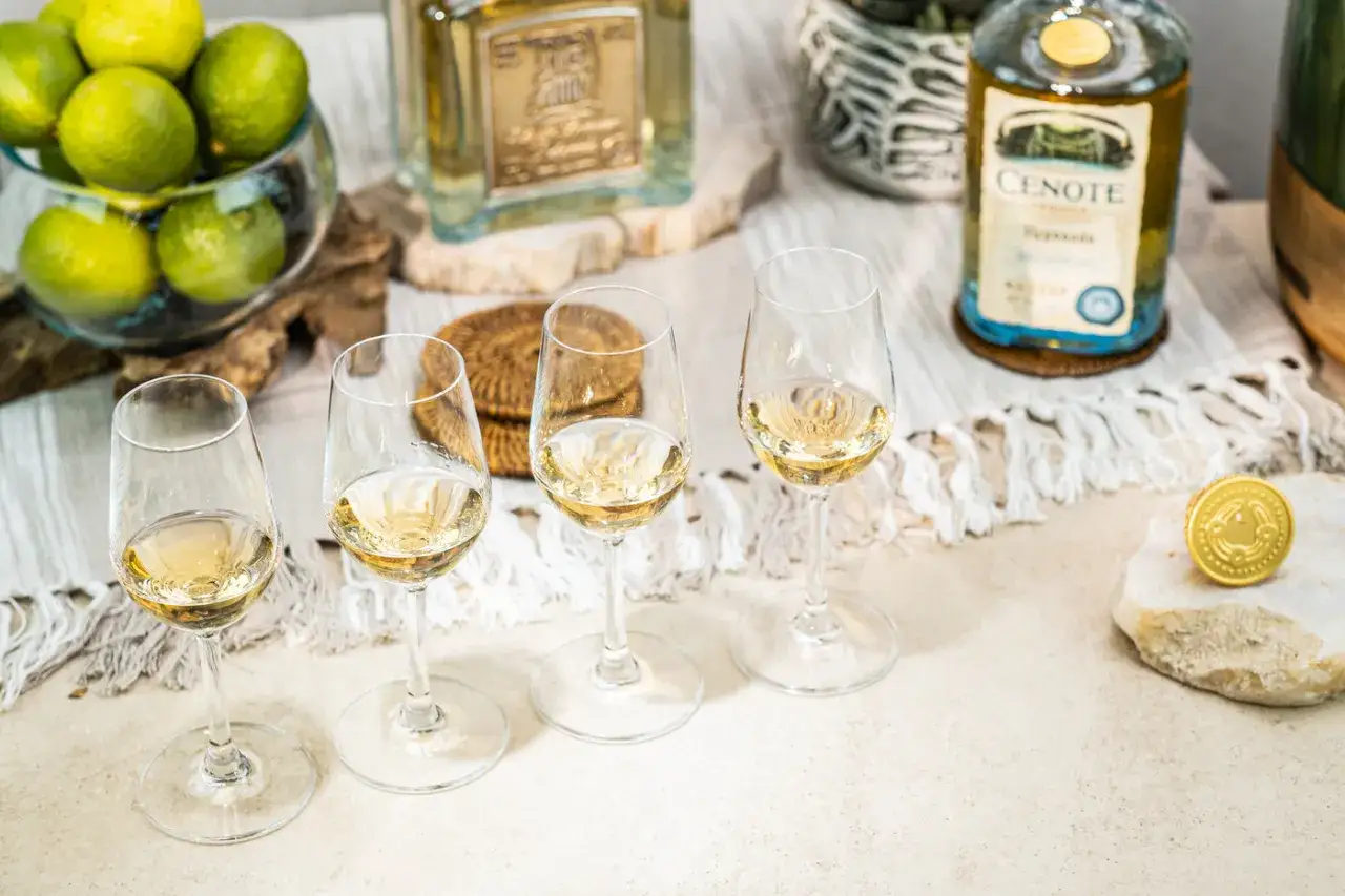 Vier Gl&auml;ser mit goldgelbem Tequila stehen bereit zum Tequila trinken. Eine Flasche Cenote Tequila und frische Limetten sind im Hintergrund zu sehen.