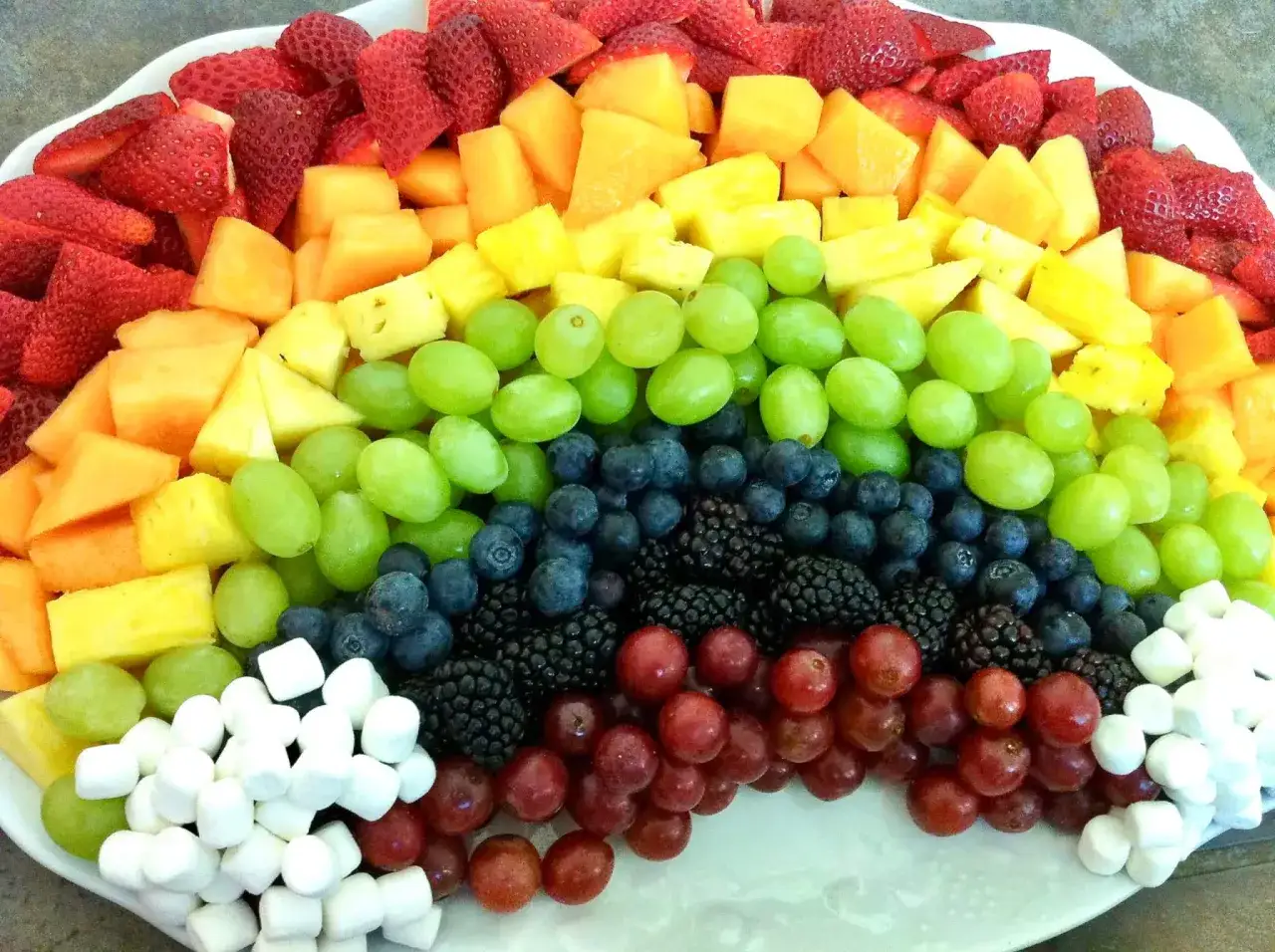 Bunter Obstteller als Regenbogen f&uuml;r Kinder: Erdbeeren, Melone, Ananas, gr&uuml;ne und rote Trauben, Blaubeeren, Brombeeren und Marshmallows.