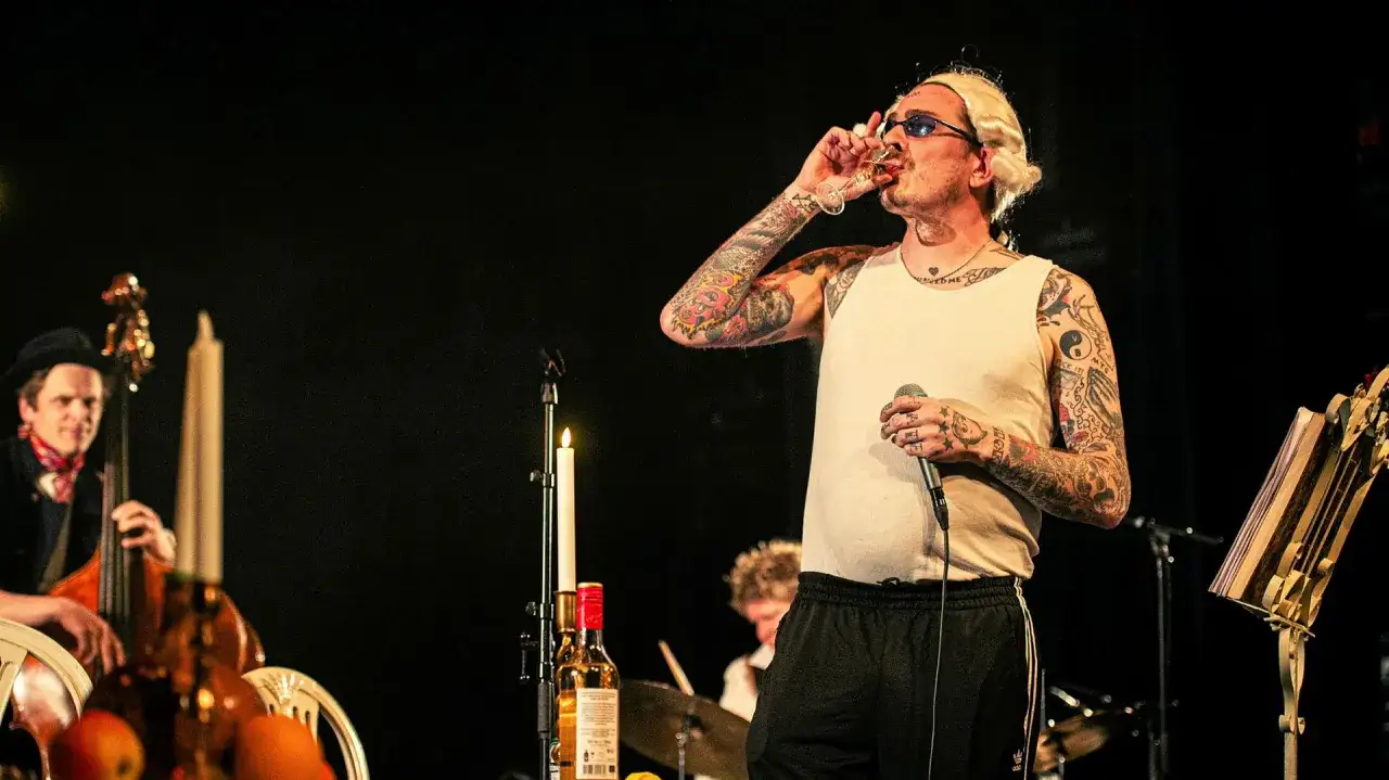 En tatuerad artist med blont hår dricker ur ett glas på scenen, med en kontrabasist och trummis i bakgrunden. En påminnelse om stockholm roots.