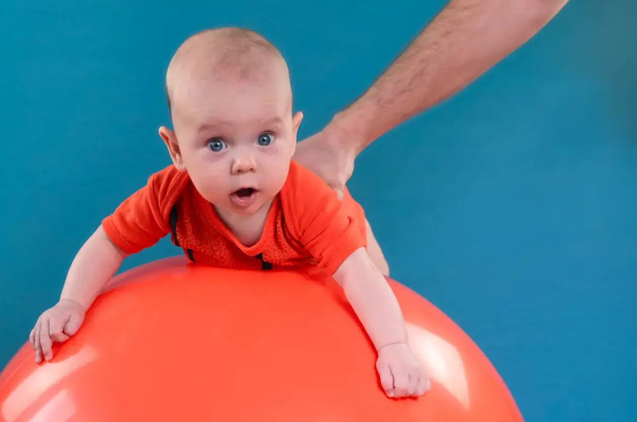 Beb&eacute; boca abajo sobre una pelota naranja, con una mano adulta sujetando su espalda. Es importante sujetar la cabeza a los beb&eacute;s para su desarrollo.