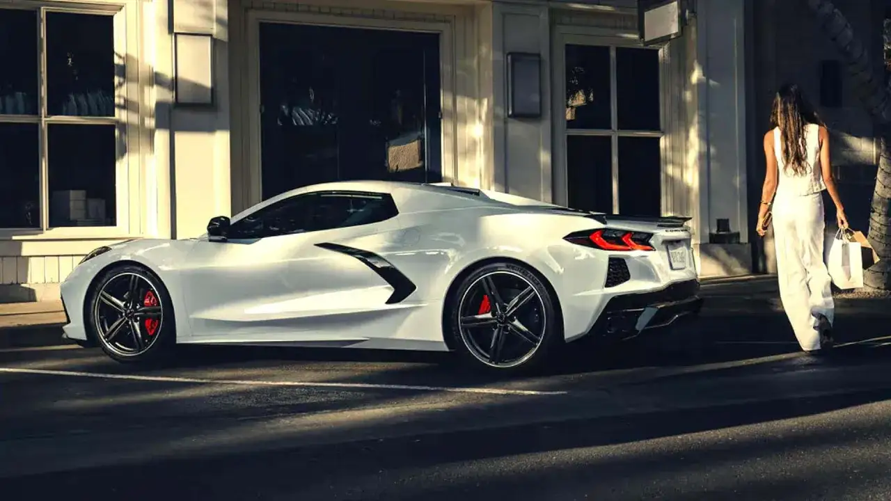 Biały Chevrolet Corvette Stingray z czerwonymi zaciskami hamulcowymi, obok eleganckiej kobiety w białym stroju, która idzie z torbami na zakupy.