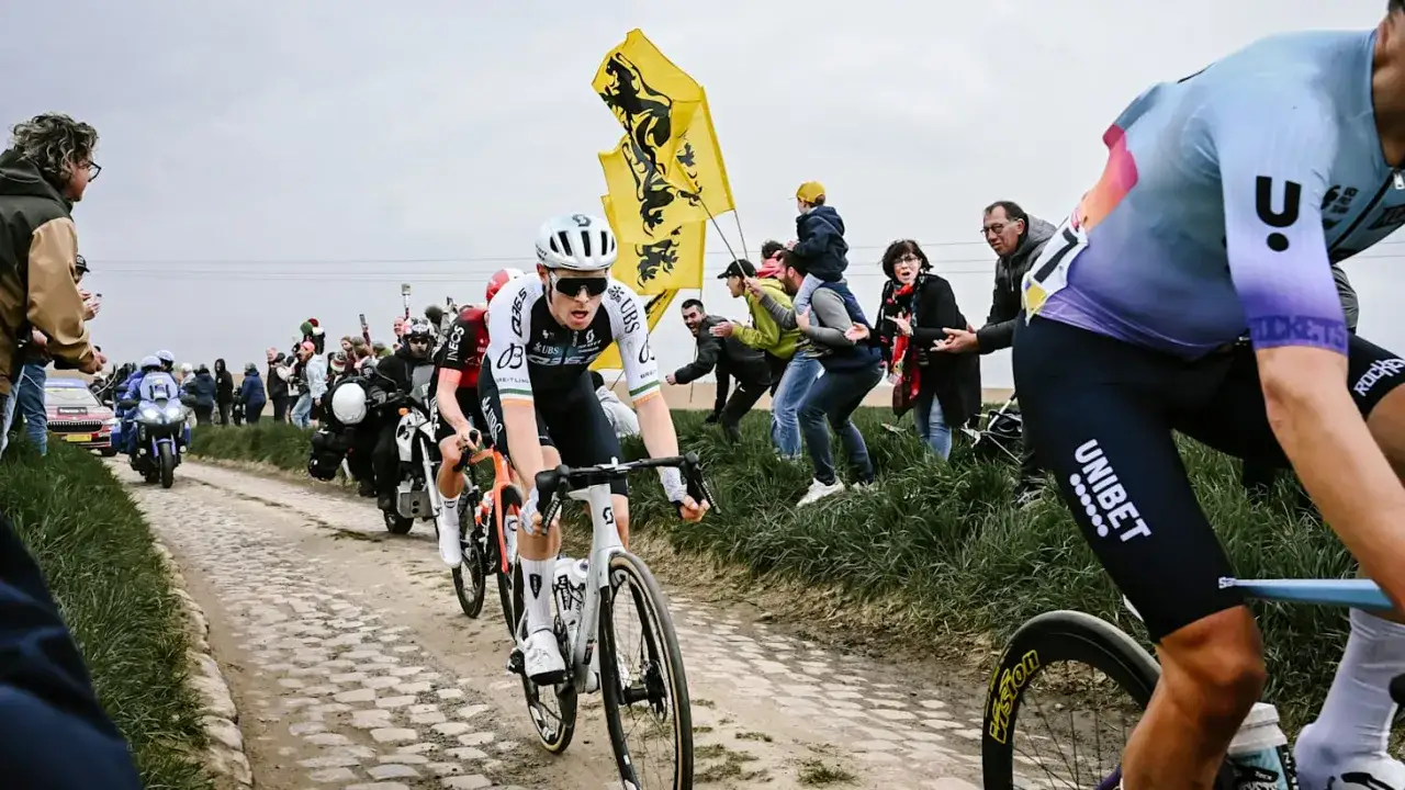 Radrennen Paris-Roubaix: Radfahrer k&auml;mpfen sich &uuml;ber Kopfsteinpflaster, w&auml;hrend Zuschauer mit Fahnen anfeuern.