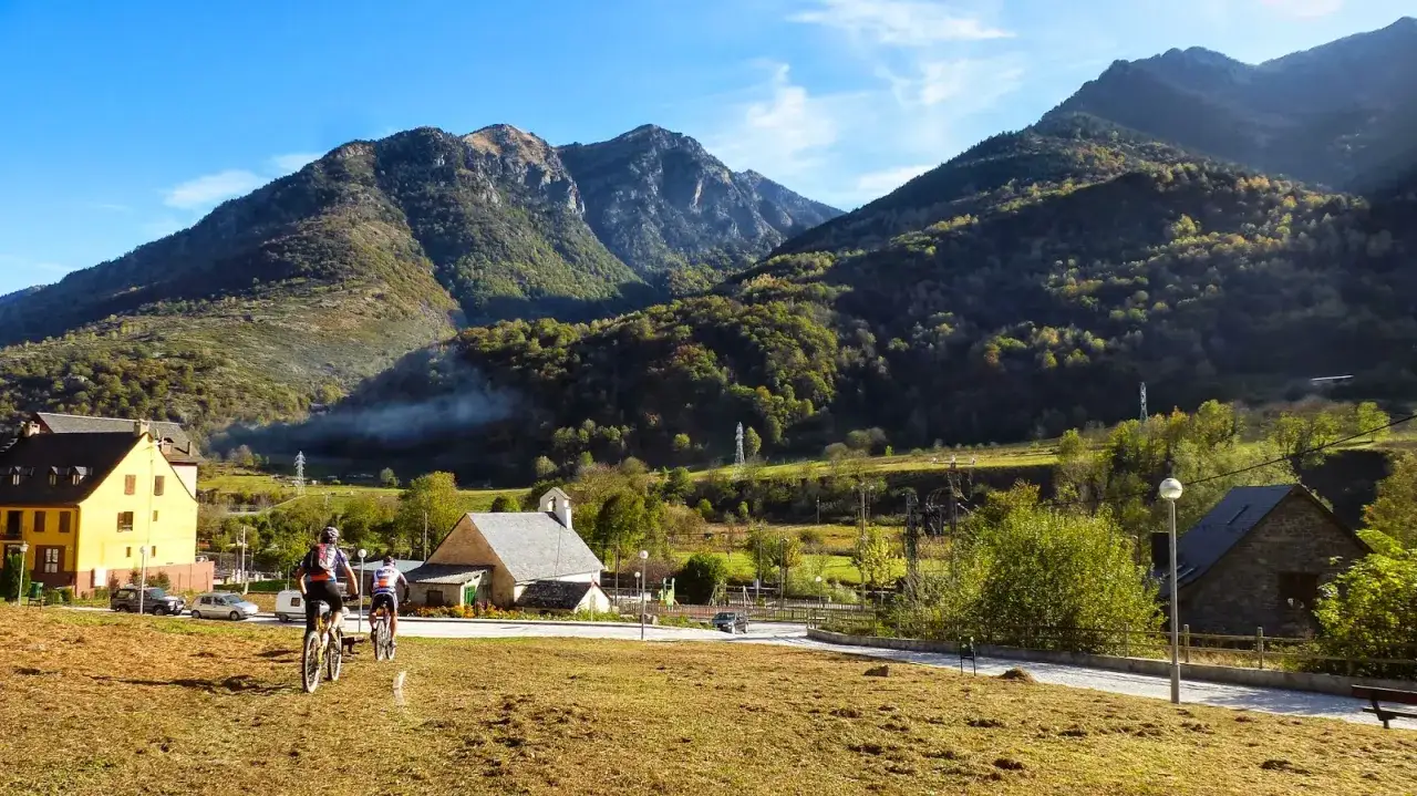 Ciclistas pedalean por un pueblo pirenaico con monta&ntilde;as verdes y niebla matutina. El paisaje de Bassa d'Arres es id&iacute;lico.