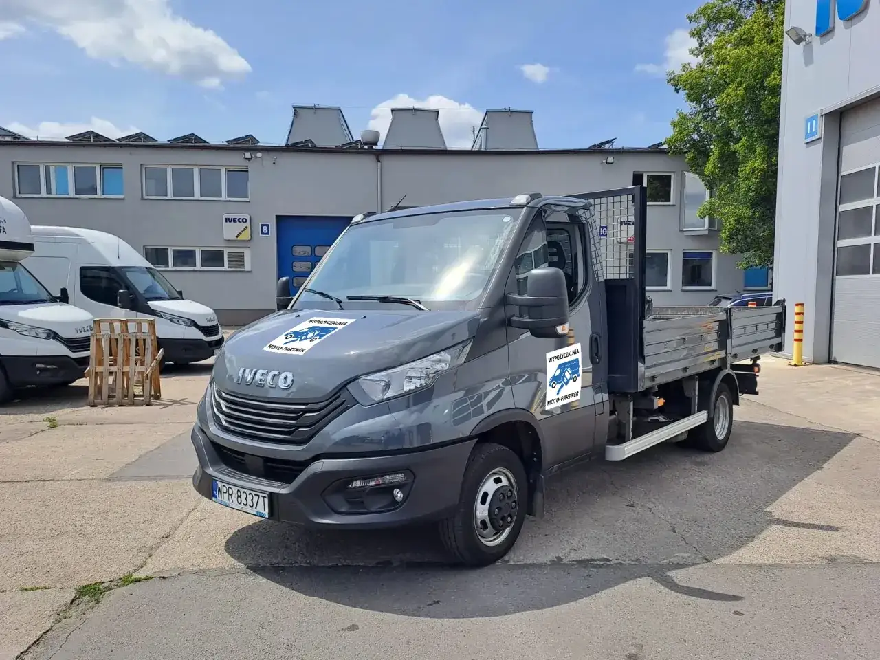 Szary Iveco z otwartą skrzynią ładunkową, idealny do wynajem wywrotki do 3,5 tony. Na drzwiach logo 