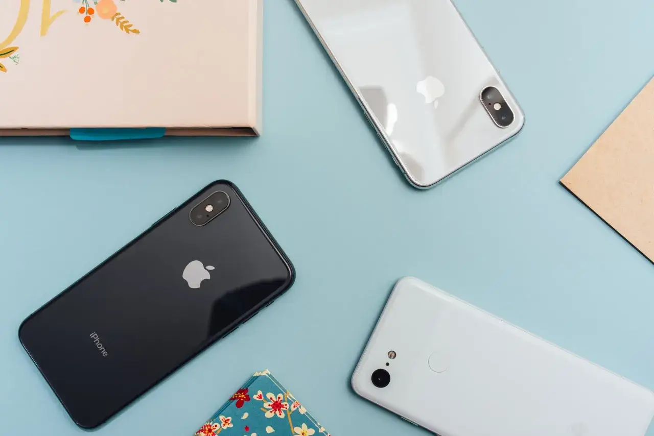Tv&aring; iPhones och en Android-telefon ligger utspridda p&aring; ett ljusbl&aring;tt underlag, bredvid en anteckningsbok och ett kuvert.