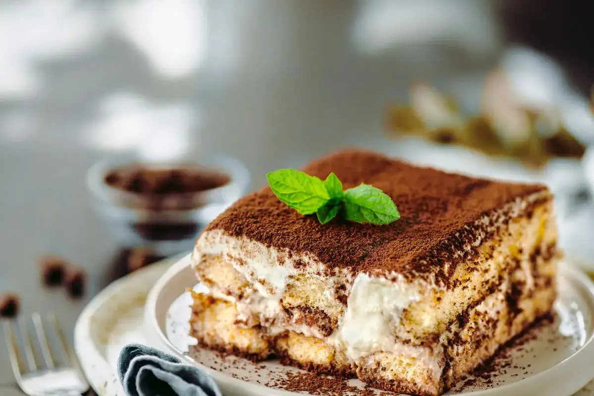 Una fetta di ricetta tiramis&ugrave; classico, con savoiardi, crema e cacao, guarnita da una fogliolina di menta.