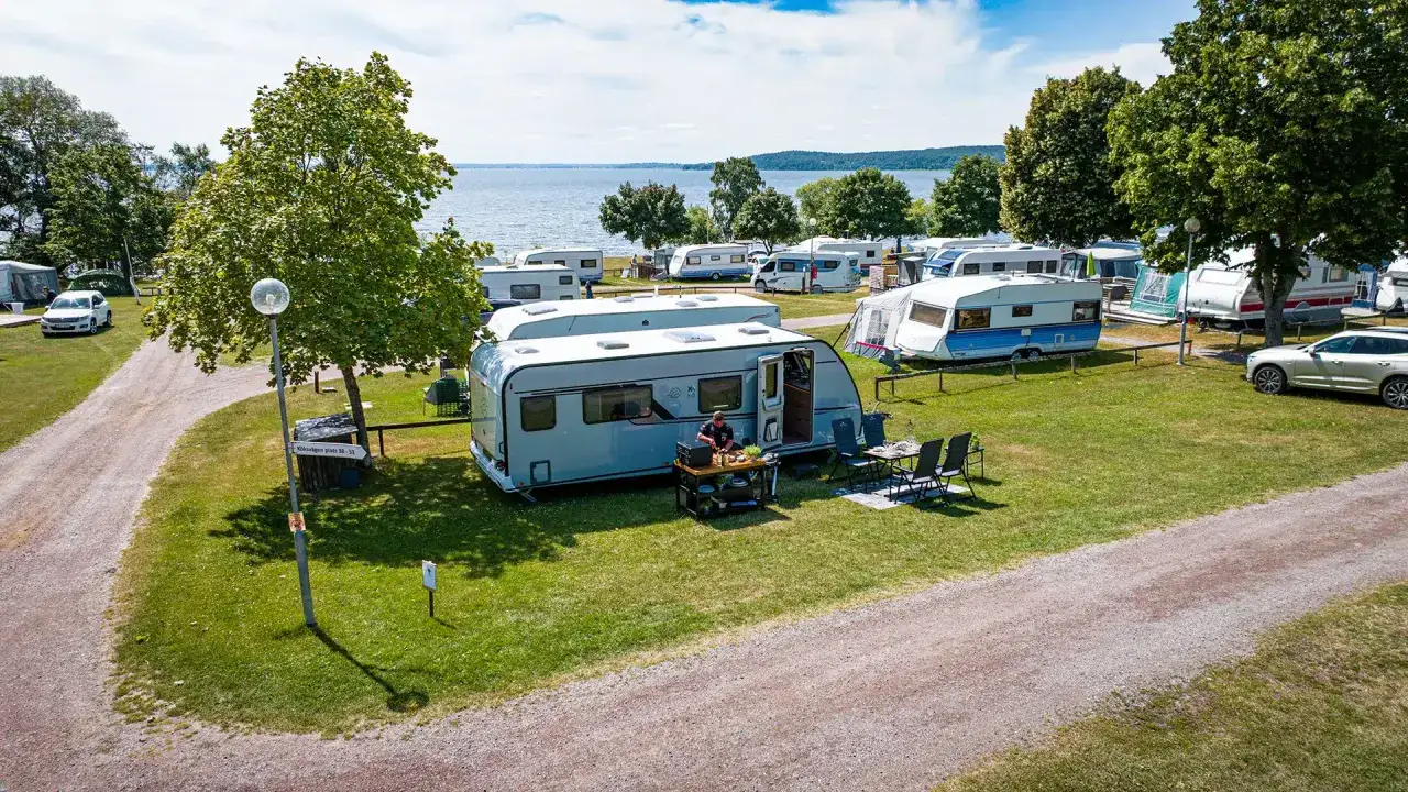 En campingplats vid en sjö i Sverige, med husvagnar uppställda på gräset. En person lagar mat utomhus vid en husvagn.