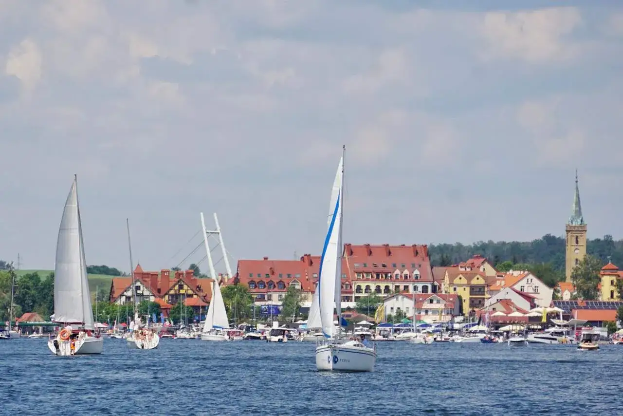 Żaglówki na jeziorze, w tle malownicze miasteczko z czerwonymi dachami. Idealne miejsce na wyjazd na Mazury.