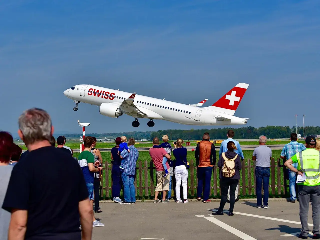 Eine Gruppe von Menschen steht am Zaun und schaut zu, wie eine Swiss-Maschine abhebt. Sie sind begeistert vom Flugzeuge fotografieren.