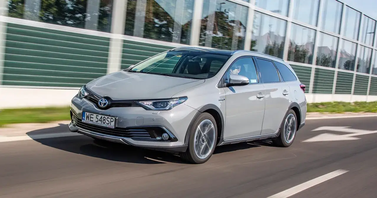Szary Toyota Auris Hybrid kombi jedzie po drodze, odbijając się w szklanej ścianie.