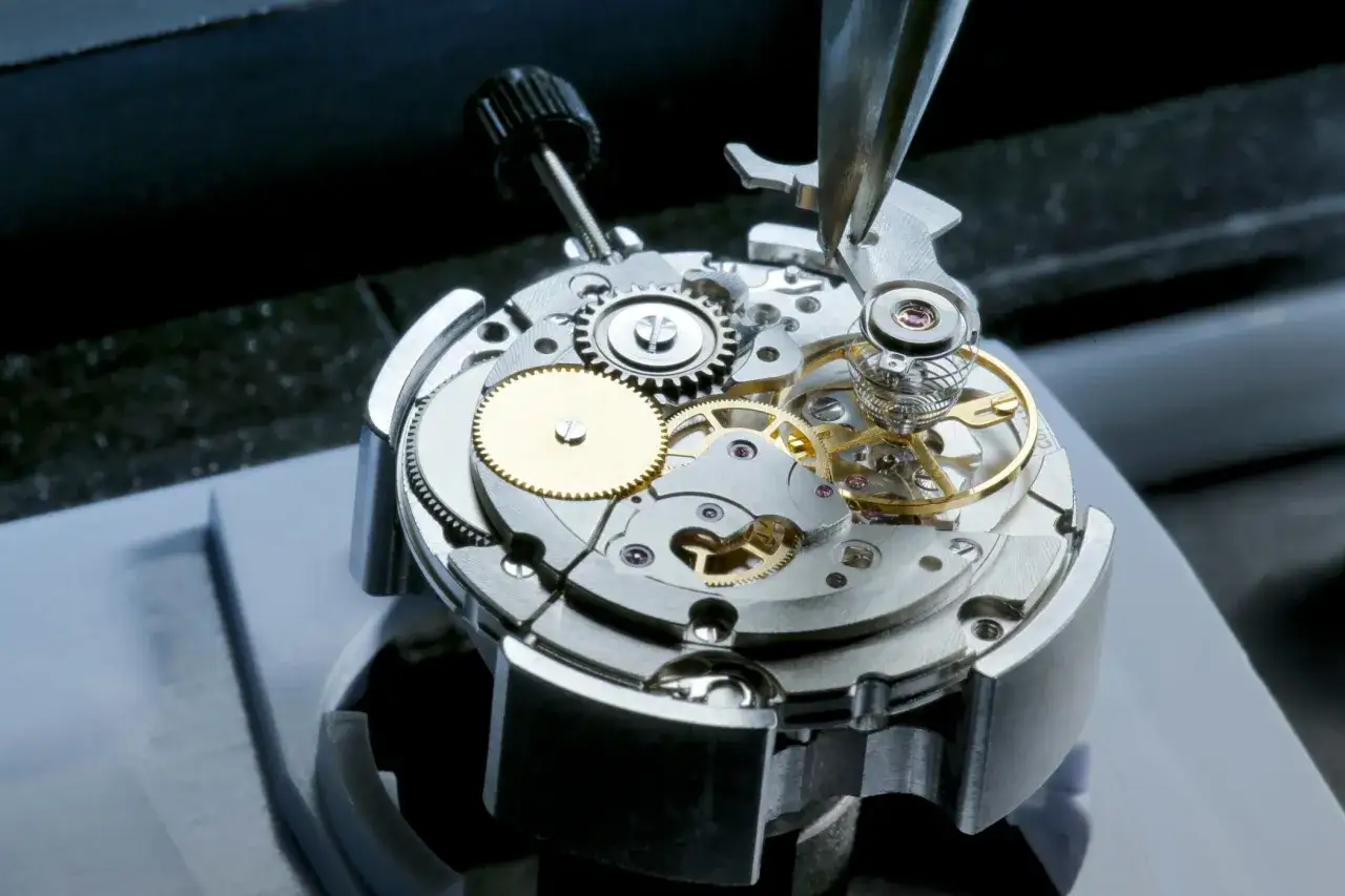 Mecanismo de reloj Powermatic 80 en montaje, con engranajes dorados y plateados, y una pinza ajustando el volante.