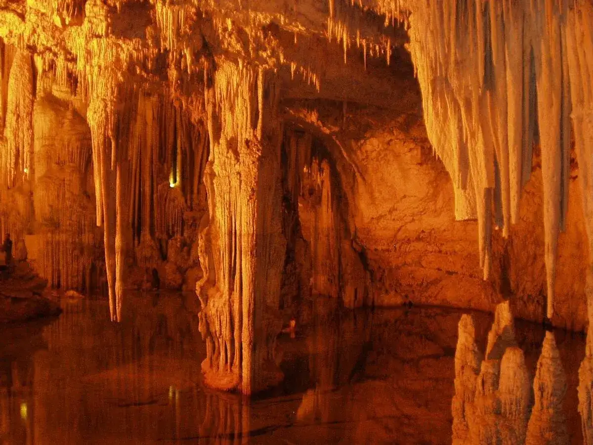 Interior de la cueva Rei Cintolo, con estalactitas y estalagmitas iluminadas en tonos c&aacute;lidos, reflej&aacute;ndose en el agua subterr&aacute;nea.