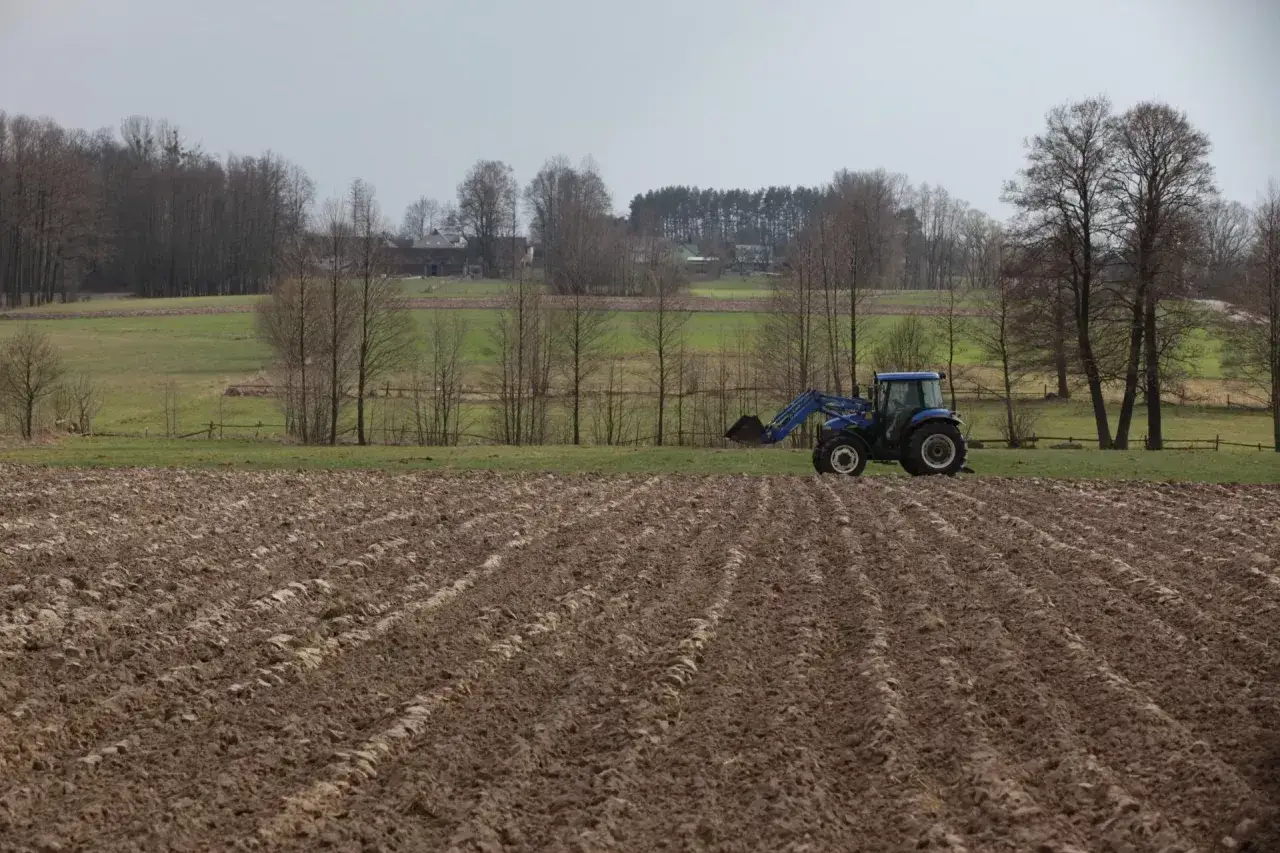 Niebieski traktor na zaoranym polu. W tle domy i drzewa. Myśląc o zakupie działki rolnej, warto pamiętać o podatku.