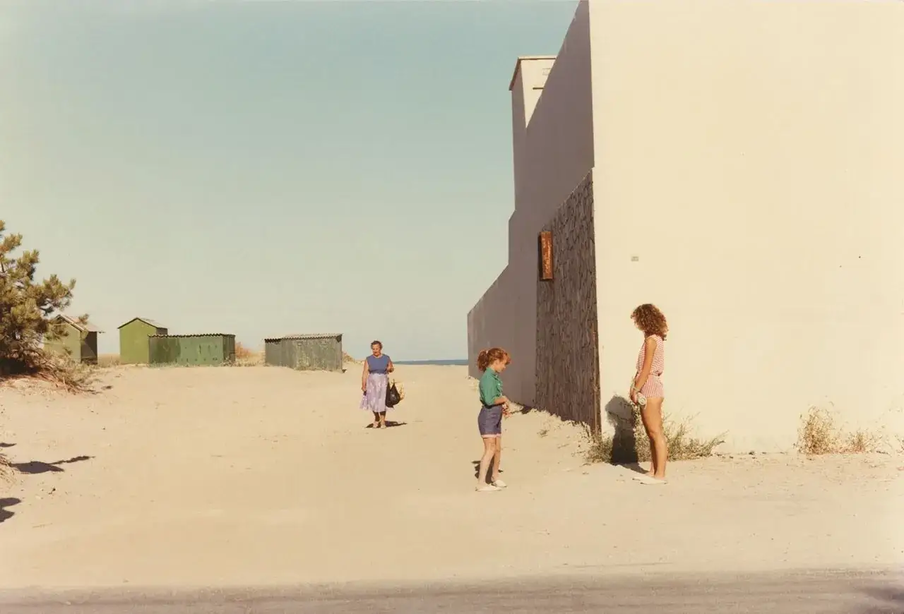 Luigi Ghirri foto: due bambine e una donna camminano su una spiaggia sabbiosa, con piccole cabine verdi sullo sfondo e un muro bianco moderno.