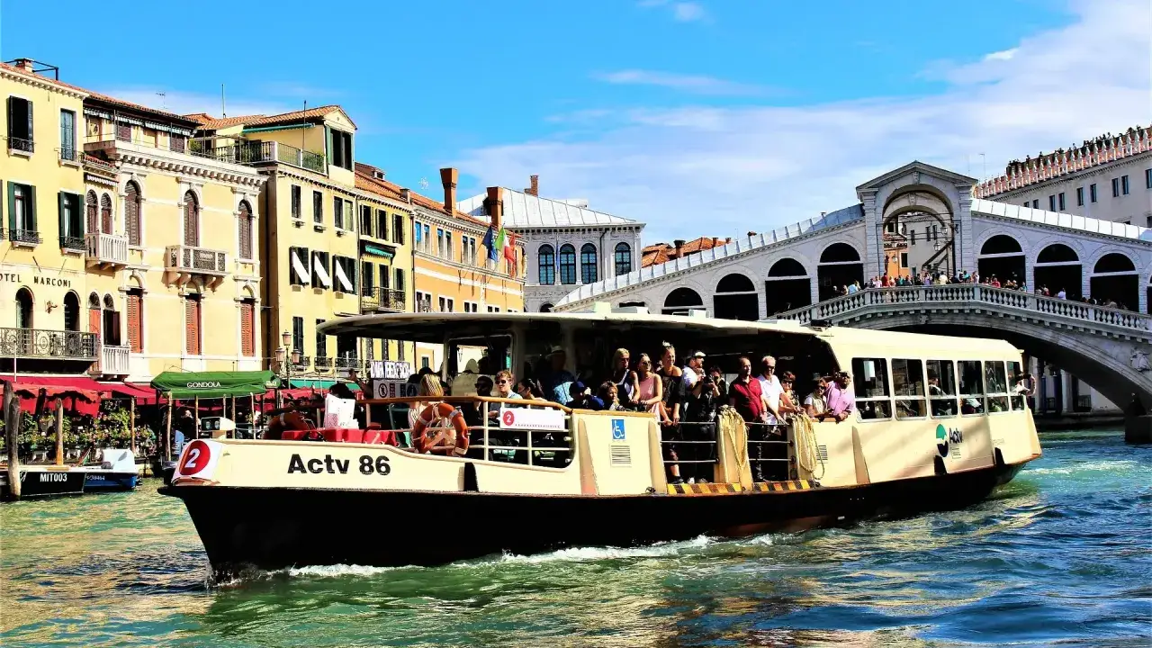 Barco lleno de turistas navega por el Gran Canal de Venecia, pasando bajo el Puente de Rialto. Un día perfecto para explorar la **Venezia unica city pass**.