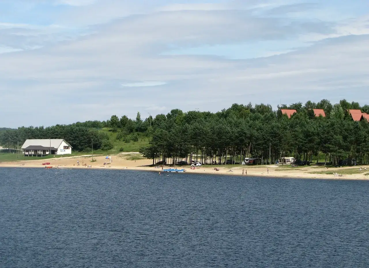 Słoneczny dzień nad jeziorem Chańcza. Plaża pełna ludzi, domki letniskowe i las tworzą malowniczy krajobraz.
