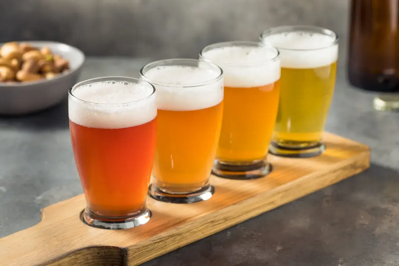 Vier Gläser mit Bier, von hellrot bis goldgelb, auf einem Holzbrett. Ein Schälchen mit Nüssen im Hintergrund. Ein Bier mit Gin-Beeren-Geschmack könnte hier gut passen.
