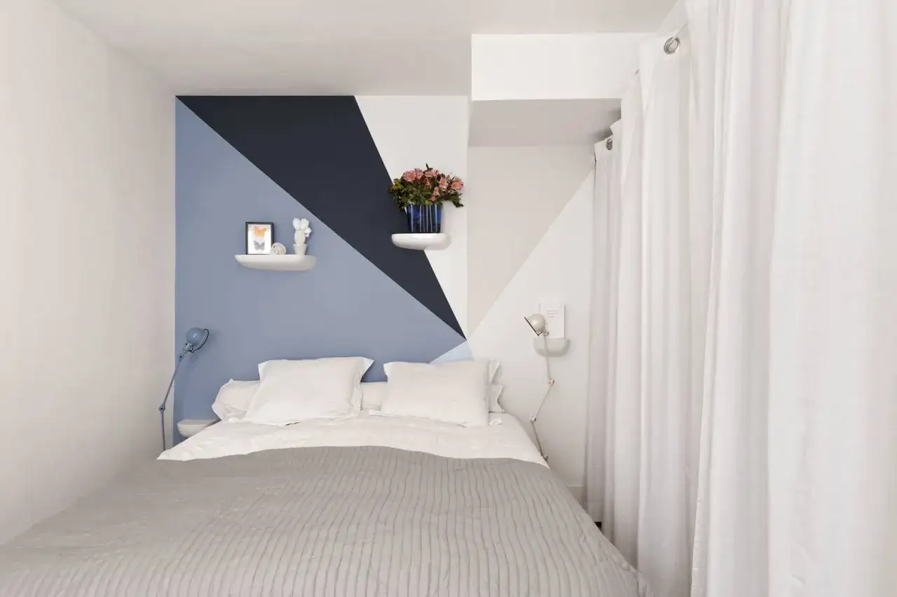 Un lit douillet sous un mur de couleur chambre aux formes géométriques bleues et blanches. Des étagères minimalistes décorent le mur.