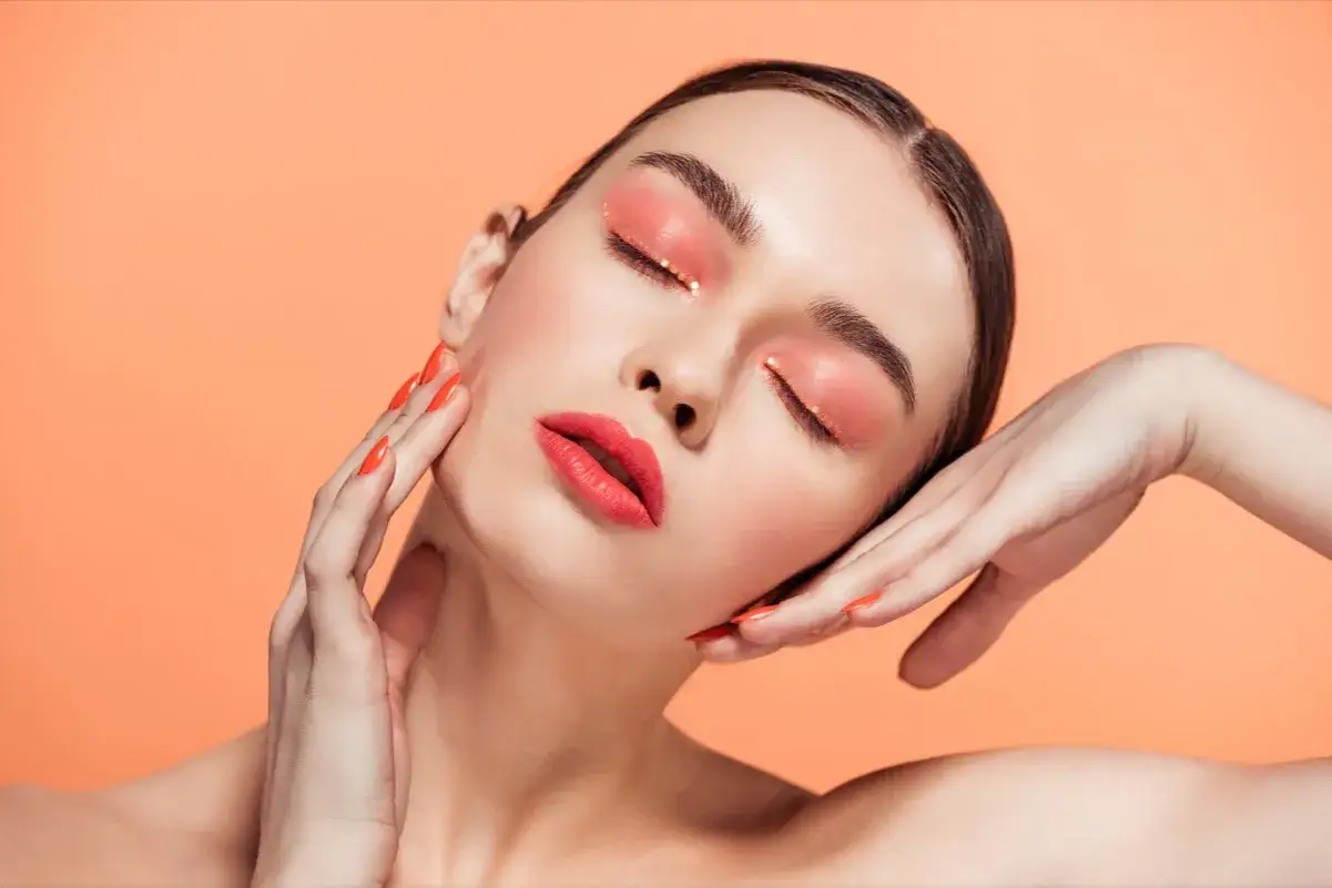 Viso di donna con trucco occhi e labbra sui toni corallo e oro, unghie arancioni. Un'ode alla **primavera soft palette**.