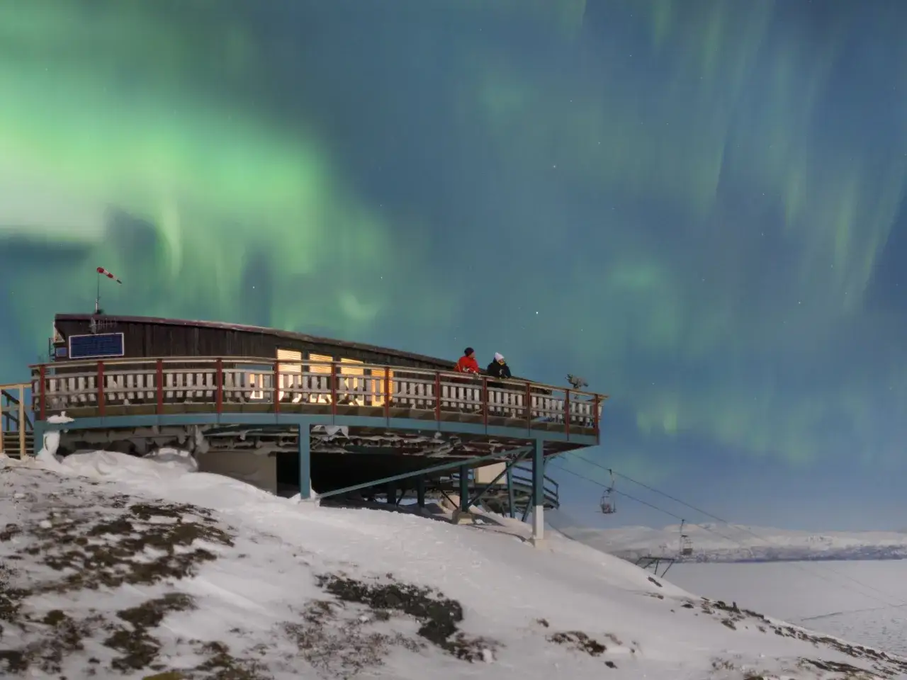 Tv&aring; personer ser p&aring; ett magiskt abisko norrsken fr&aring;n en byggnad p&aring; en sn&ouml;t&auml;ckt kulle.